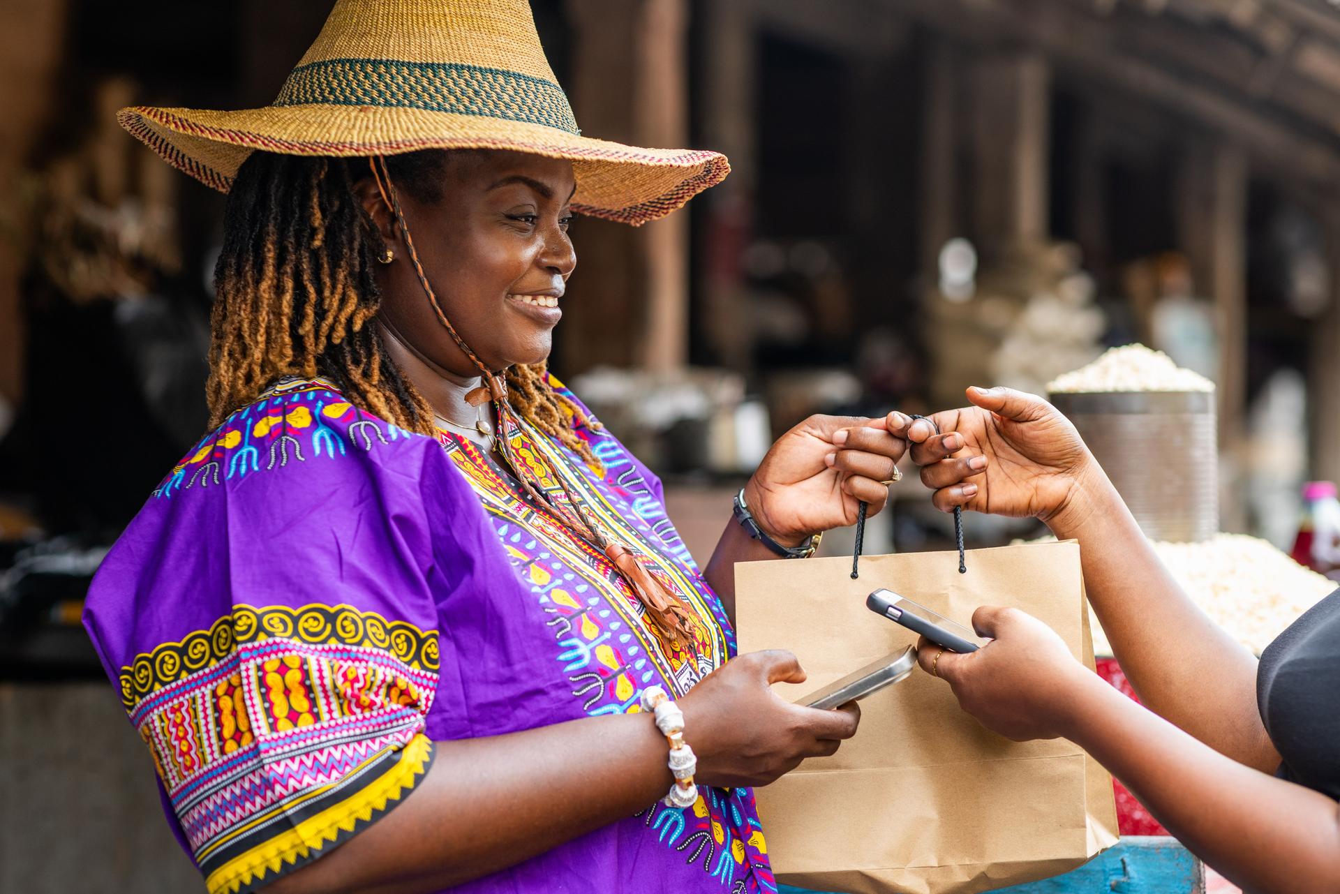 Small business owner, SME. Local traditional African market woman in a purple dashiki and straw hat receives contactless mobile money phone payment transactions at her vibrant outdoor market stall.